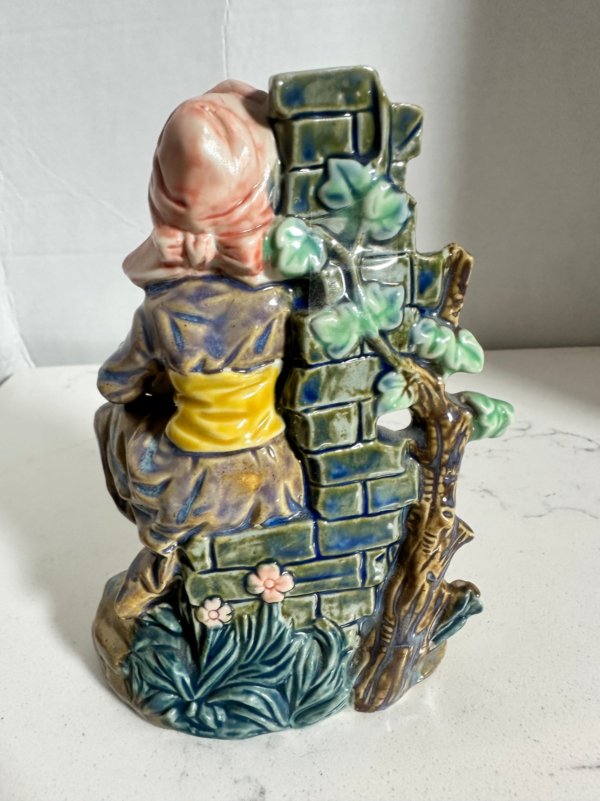 The Collected Chapter vintage ceramic fisherwoman figurine in a pink bonnet leaning against a detailed stone wall.