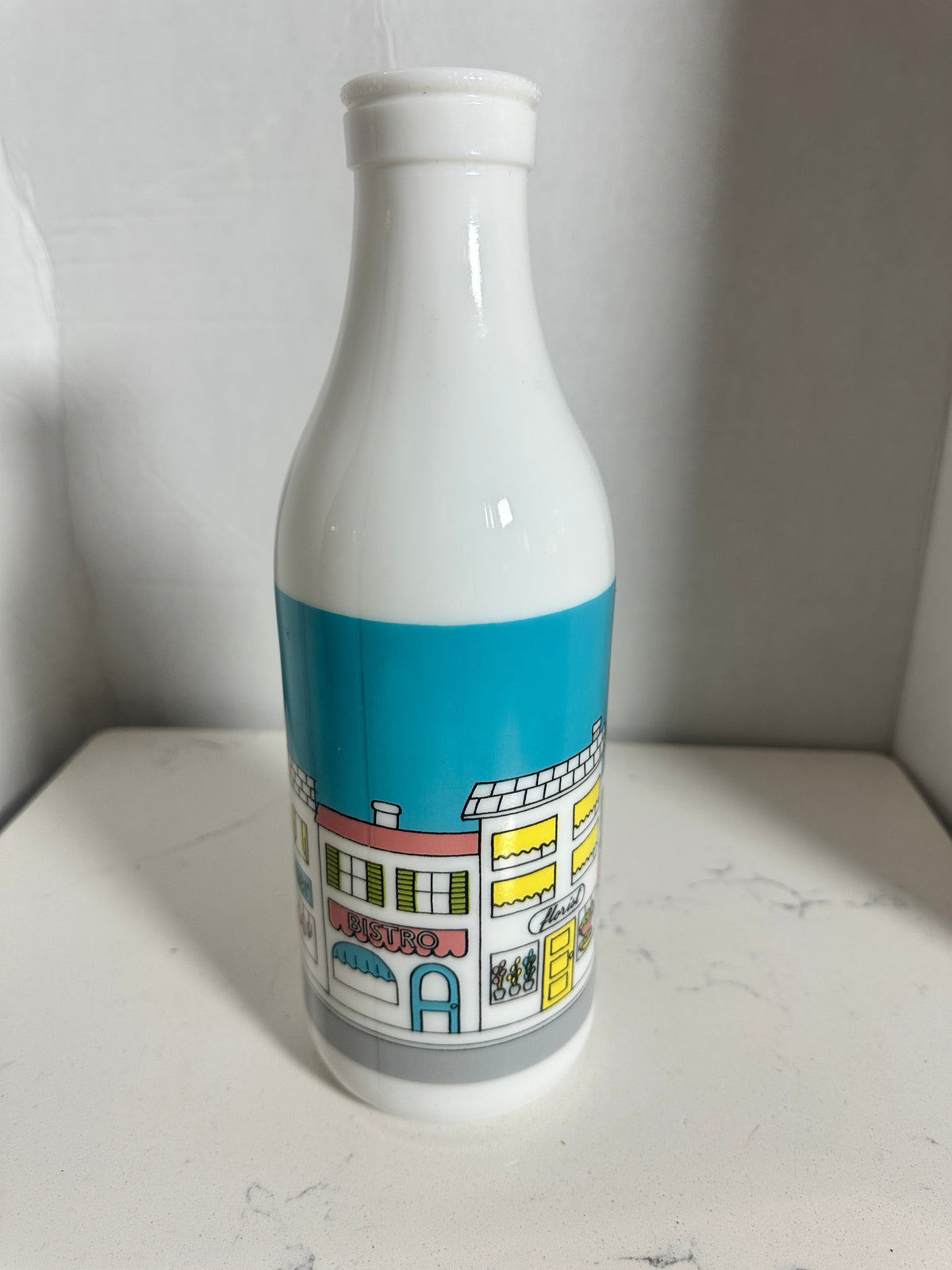 The Collected Chapter vintage white milk glass bottle vase featuring a colorful illustrated street scene storefront.